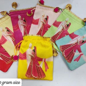 May include: A collection of six drawstring potli bags in red, yellow, pink, light pink, lime green, and light blue. Each bag features a printed illustration of a couple in traditional attire, gold trim, and pink pom-pom tassels. Size 7x9 inches.