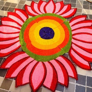 Op de afbeelding: Een kleurrijk, cirkelvormig tapijt met een bloemenontwerp. Het tapijt heeft een centrale cirkel met concentrische ringen van geel, oranje en groen, omringd door rood-roze bloemblaadjes. Het tapijt ligt op een geblokte tegelvloer.