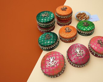 May include: A collection of colorful, round, decorative boxes in shades of green, orange, and pink. Each box is adorned with a gold-colored emblem and a pearl-like trim. The boxes are stacked and arranged on a two-tone background.