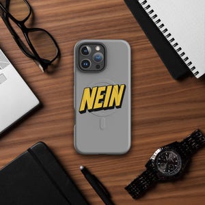 Könnte beinhalten: Eine mattgraue Smartphone-Hülle mit dem Wort NEIN in fetten gelben Großbuchstaben und schwarzem Schlagschatten. Die Hülle hat ein kreisförmiges magnetisches Design auf der Rückseite und eine Aussparung für ein Triple-Kamera-System oben links.