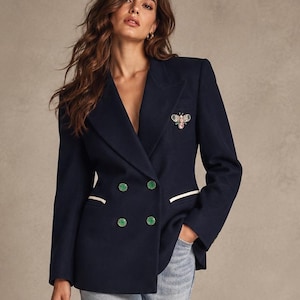 Peut inclure: Un blazer bleu marine à double boutonnage avec quatre boutons verts, des bordures de poches blanches et un petit écusson brodé en forme d'abeille sur la poitrine, porté avec un jean bleu clair et des chaussures noires à bout pointu.