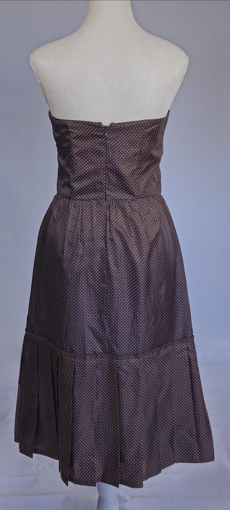 May include: A strapless, knee-length dress in a dark brown fabric with a white polka dot pattern. The dress features a fitted bodice and a pleated skirt. The dress is displayed on a mannequin.