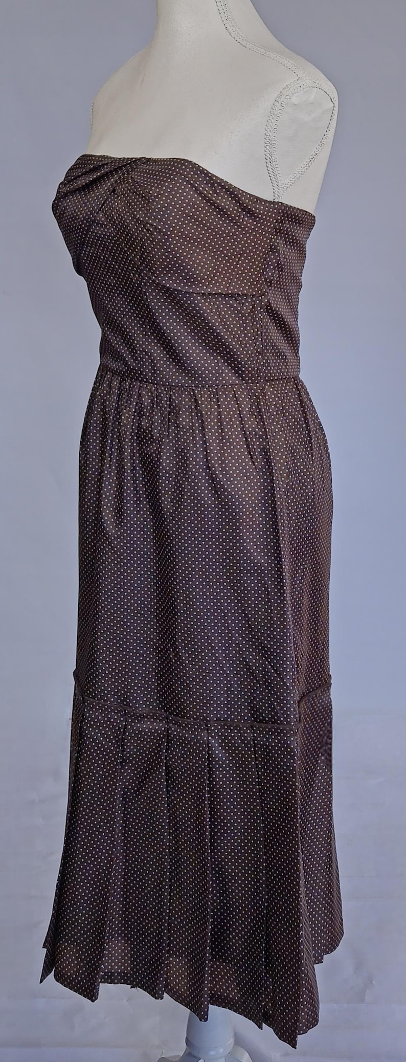May include: A brown strapless dress with a white polka dot pattern. The dress features a fitted bodice, a gathered waist, and a pleated skirt. The dress is displayed on a mannequin.