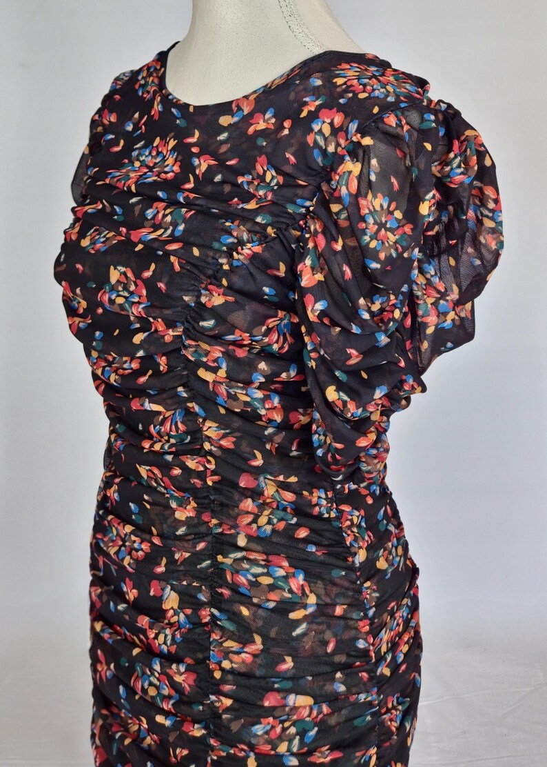 May include: Black dress with a floral pattern in red, yellow, blue, and green. The dress features a round neckline and short, ruffled sleeves. The fabric is gathered throughout the bodice and skirt.