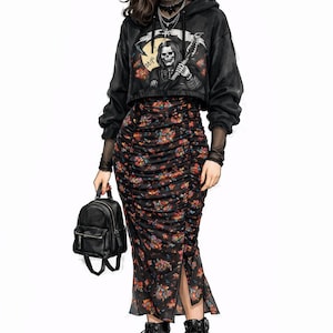 May include: A black cropped hoodie with a Grim Reaper graphic, paired with a floral print ruched skirt and black combat boots. A small black backpack is held in the hand. The outfit is a blend of edgy and feminine styles.