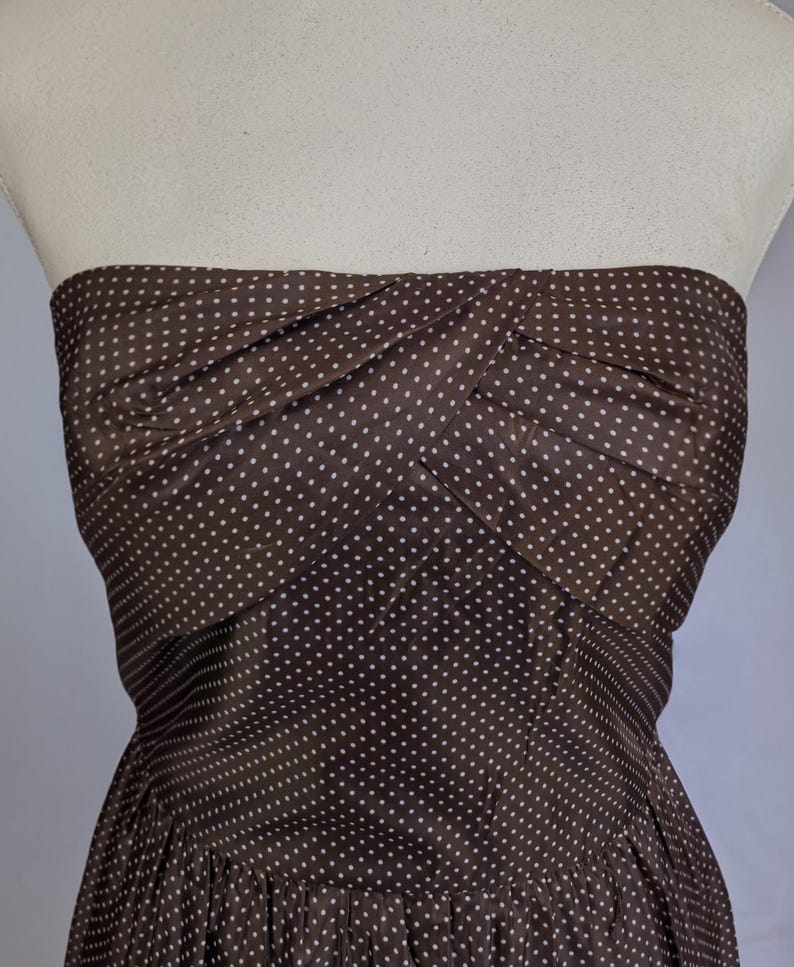 May include: A strapless brown dress with white polka dots. The bodice has a twisted design, and the skirt is gathered. The dress is displayed on a mannequin.