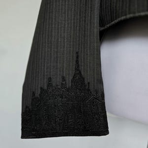 Peut inclure: Gros plan sur un tissu à fines rayures gris foncé avec un motif de paysage urbain brodé en noir le long du bord. Le tissu semble faire partie d'un vêtement, peut-être une veste ou un blazer.