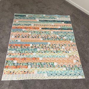 May include: A colorful quilt with a patchwork design. The quilt features horizontal stripes in various colors, including peach, teal, and white, with floral and geometric patterns. The quilt is made of fabric and is laid out on a gray carpet.