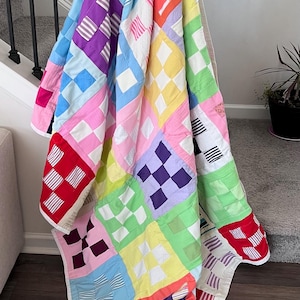 May include: A vibrant patchwork quilt with a range of geometric designs and a white border. The quilt displays squares in shades of red, pink, purple, blue, yellow, and green. The quilt is draped over a staircase.