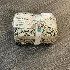 Peut inclure: Un paquet de tissu emballé dans du plastique transparent et noué avec un ruban. Le tissu présente un motif floral noir sur fond crème. Le ruban est blanc avec le mot "moda" imprimé dessus.