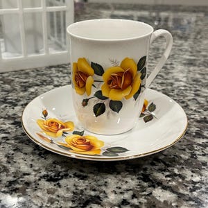 May include: White teacup and saucer set with a gold rim and yellow rose floral design. The teacup has a handle and the saucer is round. The set is on a granite countertop.