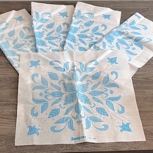 Puede incluir: Cinco cuadrados de tela blanca con un diseño floral azul claro. El diseño incluye hojas, flores y detalles en forma de estrella. La tela está dispuesta sobre una superficie de madera. El texto "Janlyn" es visible en uno de los cuadrados.