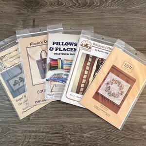May include: Several craft pattern packets in clear plastic sleeves are arranged on a wooden surface. The packets feature various designs, including floral wreaths, pillows, and coasters. The packets have text such as "WEE WELCOME" and "Backyard Friends."