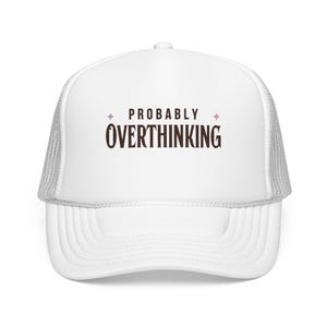 May include: A white trucker hat with a mesh back and a solid front panel. The front features the text "PROBABLY OVERTHINKING" in a dark brown serif font, flanked by two small pink four-pointed star icons on either side of the word "PROBABLY".