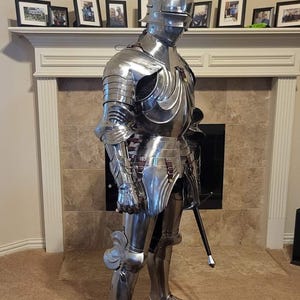Puede incluir: Una armadura completa de acero pulido, que incluye un casco, peto, protección para brazos y piernas y cubiertas para los pies. La armadura se exhibe en interiores, de pie. Una espada negra se sostiene a un lado. El fondo incluye una chimenea y un reloj.