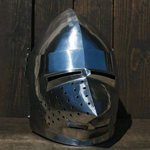 Puede incluir: Un casco medieval pulido de color plateado, con una parte superior puntiaguda y una visera con ranuras horizontales para los ojos y numerosos orificios de ventilación pequeños. La superficie del casco refleja el entorno, creando una apariencia brillante. El diseño recuerda a la armadura histórica.