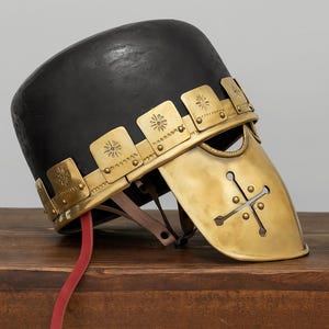 Puede incluir: Un casco negro y dorado con un diseño decorativo. El casco tiene una parte superior redondeada y una protección facial dorada con un diseño de cruz. El casco tiene una correa de cuero roja colgando del costado.