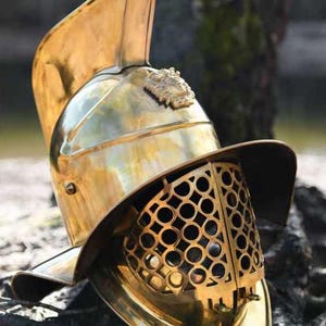 Puede incluir: Un casco de gladiador de latón pulido con una cresta y una protección facial prominentes. El casco presenta un diseño detallado, con un patrón circular en la protección facial y un emblema decorativo en la parte superior. Réplica histórica.