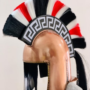 May include: A copper-colored Spartan helmet with a black, white, and red plume. The helmet features a decorative band with a Greek key pattern. The helmet has a chin strap and a polished finish.