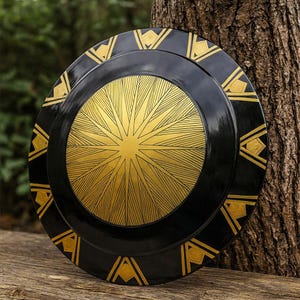 May include: A black and gold shield with a sunburst design in the center and geometric patterns around the edge. The shield is round and appears to be made of metal. The gold elements provide a striking contrast against the black.