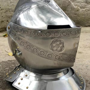 May include: A polished silver metal helmet with detailed floral and geometric patterns. The helmet includes a visor and a neck guard with decorative rivets. The design evokes a medieval or historical aesthetic.