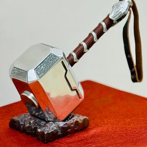 May include: A silver-colored hammer with intricate designs on the head and handle. The handle is wrapped in brown leather and has a leather strap. The hammer is resting on a dark brown, textured base, set against a red background.