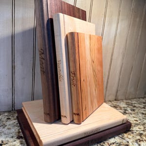 May include: A collection of wooden cutting boards in various sizes and wood grains. The boards are engraved with the words "Meat", "Vegetables", and "Fruit". The boards are stacked and arranged to stand upright, showcasing their different wood tones.
