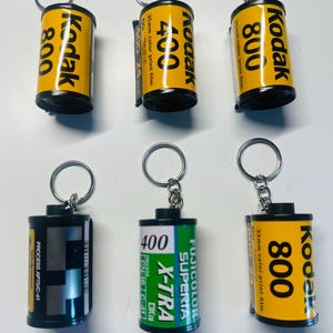 Pode incluir: Seis chaveiros feitos de recipientes de filme de 35 mm reaproveitados. Os chaveiros apresentam a marca Kodak em amarelo e preto, com etiquetas "800" e "400". Um chaveiro é verde com o texto "Fujicolor Superia X-TRA". Cada chaveiro tem um porta-chaves prateado.