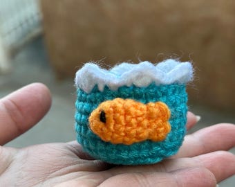 May include: A small, crocheted fishbowl in teal with a white wave trim. An orange fish is appliqued on the side. The handmade item is held in a hand.