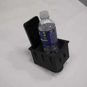 May include: A black plastic cup holder with an integrated vertical slot for a smartphone. A clear plastic water bottle with a blue and white label is placed inside the circular cup holder section of the rectangular black accessory.