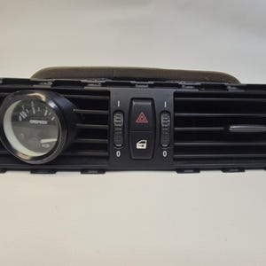 Op de afbeelding: Zwarte auto dashboard luchtventilatie met een meter en bedieningsknoppen. De meter heeft een witte wijzerplaat met zwarte markeringen. De ventilatie heeft horizontale lamellen en een rechthoekige vorm.