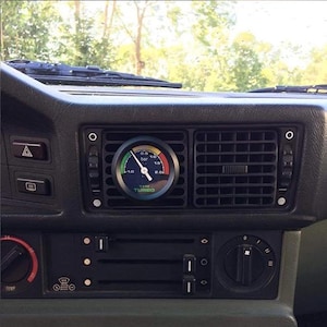May include: A car dashboard featuring a turbo gauge mounted in the air vent. The gauge has a black face with a white needle and a scale marked in bar. The text "TIME TURBO" is visible. The dashboard is black.