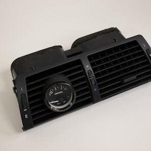 May include: Black car dashboard air vent with a round gauge in the center. The gauge reads "OIL" and "CRUISPEED". The vent has adjustable louvers and control knobs. The item is set against a white background.