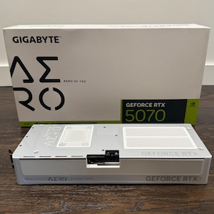 Op de afbeelding: Een witte Gigabyte Aero OC 12G grafische kaart met een zilveren metallic afwerking, voorzien van twee witte koelventilatoren en de tekst GeForce RTX op de zijkant. Daarachter staat de originele witte doos met het label Gigabyte Aero en GeForce RTX 5070.