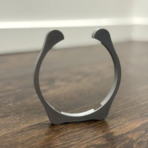 Peut inclure: Un objet circulaire gris foncé avec un design incurvé. L'objet comporte deux extensions incurvées vers le haut sur les côtés opposés. Il est fait d'un matériau texturé et repose sur une surface en bois.