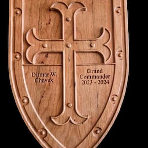 May include: A wooden shield featuring a carved cross design, with the text "Ditmar W. Chavex" and "Grand Commander 2023-2024". The shield is light brown and has a decorative border with small circular accents. The shield is likely a commemorative item.