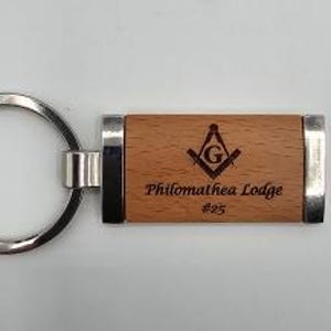 May include: A wooden keychain with a silver key ring and metal accents. The wooden portion features the text "Philomathea Lodge #25" and a Masonic symbol.