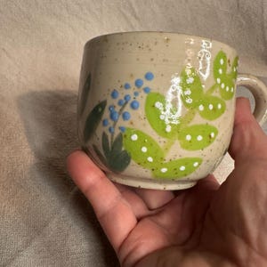 Pode incluir: Uma caneca de cer&acirc;mica com acabamento bege salpicado. A caneca apresenta folhas verdes pintadas &agrave; m&atilde;o com bolinhas brancas e cachos de bagas azuis. A caneca tem formato arredondado e uma pequena al&ccedil;a.