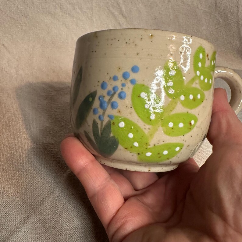 Pode incluir: Uma caneca de cer&acirc;mica com acabamento bege salpicado. A caneca apresenta folhas verdes pintadas &agrave; m&atilde;o com bolinhas brancas e cachos de bagas azuis. A caneca tem formato arredondado e uma pequena al&ccedil;a.