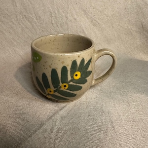 Pode incluir: Uma caneca de cer&acirc;mica bege salpicada com uma al&ccedil;a curva. A caneca apresenta um design floral verde e amarelo, com folhas verdes e detalhes circulares amarelos. A caneca est&aacute; sobre uma superf&iacute;cie de tecido de cor neutra.