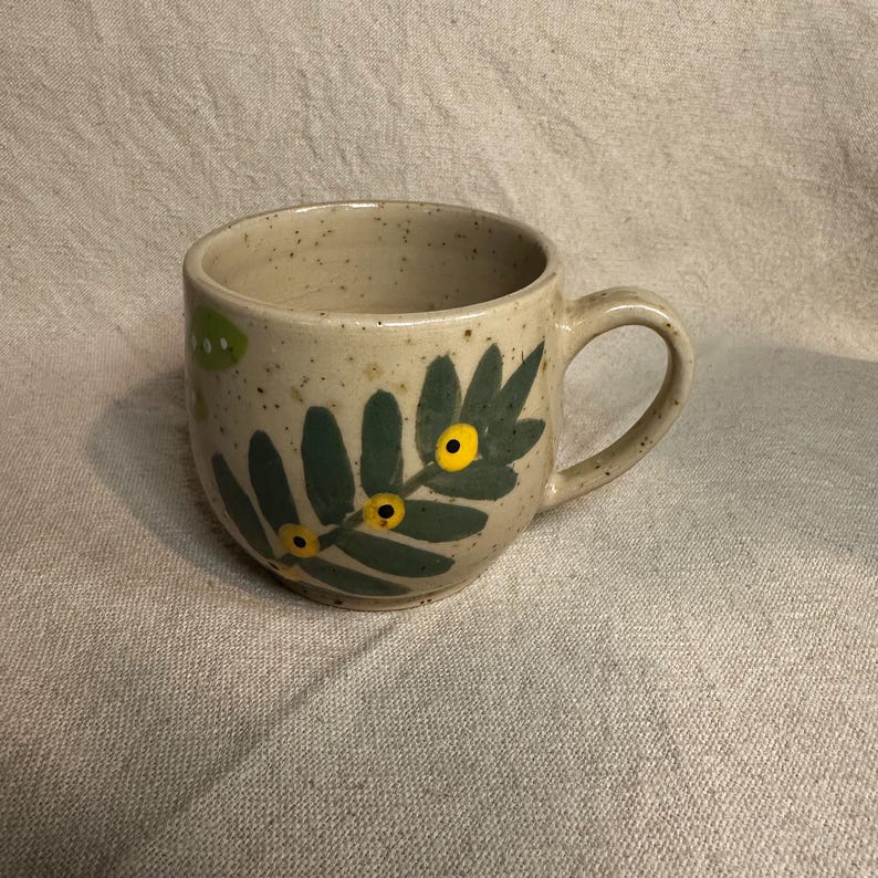 Pode incluir: Uma caneca de cer&acirc;mica bege salpicada com uma al&ccedil;a curva. A caneca apresenta um design floral verde e amarelo, com folhas verdes e detalhes circulares amarelos. A caneca est&aacute; sobre uma superf&iacute;cie de tecido de cor neutra.