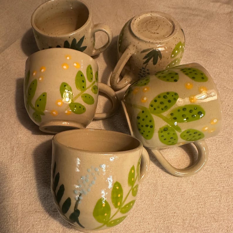 Pode incluir: Uma cole&ccedil;&atilde;o de cinco canecas de cer&acirc;mica feitas &agrave; m&atilde;o. As canecas bege apresentam desenhos de folhas verdes e pontos amarelos. Cada caneca tem uma al&ccedil;a e uma forma &uacute;nica, mostrando a arte da cer&acirc;mica.