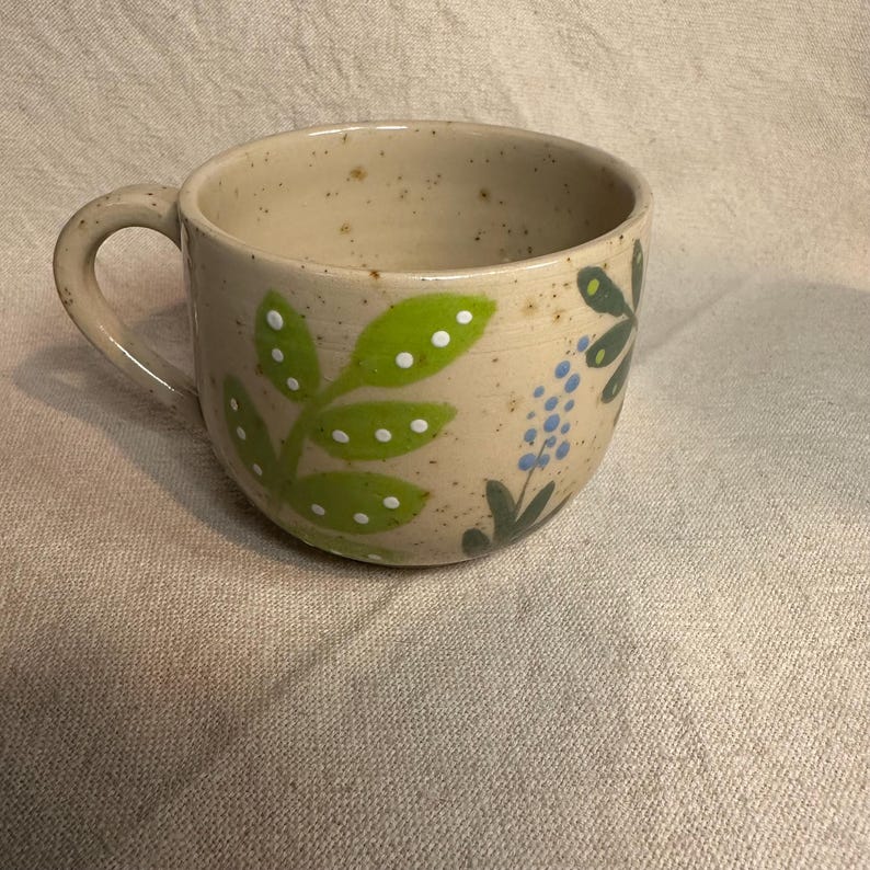 Pode incluir: Uma caneca de cer&acirc;mica bege com uma al&ccedil;a, decorada com folhas verdes com pontos brancos e detalhes florais azuis. A caneca tem uma textura salpicada e uma forma arredondada, adequada para caf&eacute; ou ch&aacute;.