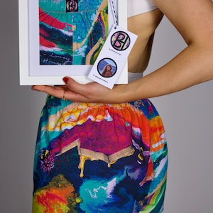 Könnte beinhalten: Ein gerahmtes Kunstwerk mit einem abstrakten Design in Türkis-, Rot- und Gelbtönen wird vor einer bunten Shorts gehalten. Die Shorts weisen ein lebendiges, mehrfarbiges Muster mit einem Wabenmuster auf. Ein Etikett mit Logo und Foto ist angebracht.