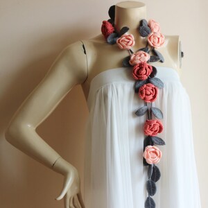 May include: A handcrafted crochet necklace featuring a garland of roses in shades of coral, peach, and gray. The floral design is accented with gray leaves, creating a unique and artistic accessory. The necklace is displayed on a mannequin.