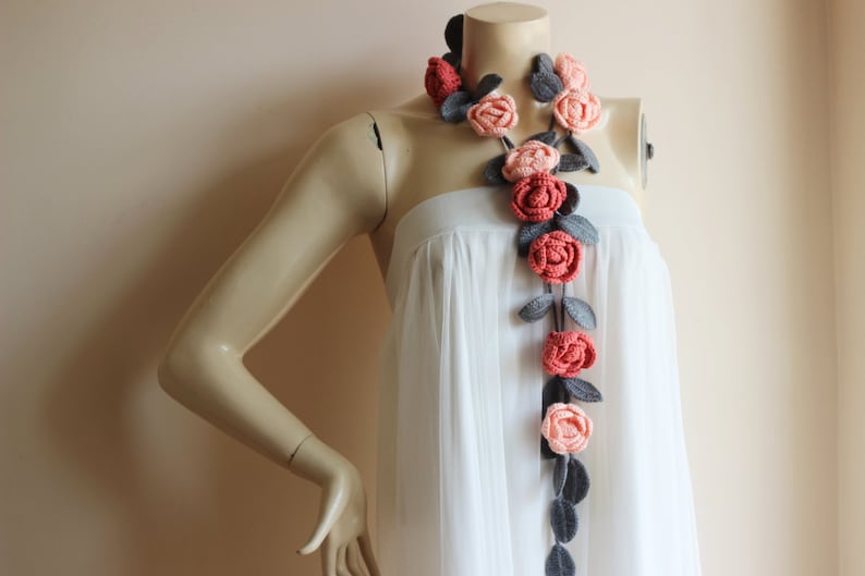May include: A handcrafted crochet necklace featuring a garland of roses in shades of coral, peach, and gray. The floral design is accented with gray leaves, creating a unique and artistic accessory. The necklace is displayed on a mannequin.