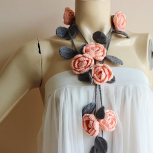 May include: A handmade crochet necklace featuring peach-colored roses and gray leaves. The floral design is arranged on a gray cord, creating a delicate and feminine accessory. The necklace is displayed on a mannequin.