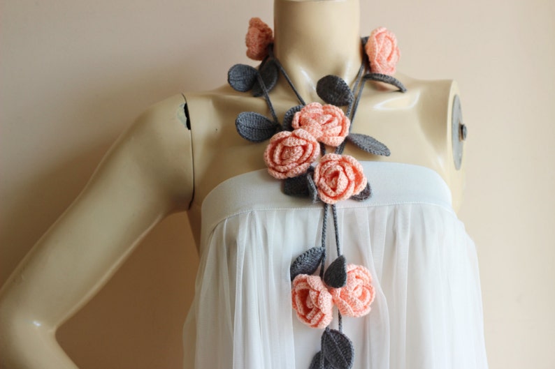 May include: A handmade crochet necklace featuring peach-colored roses and gray leaves. The floral design is arranged on a gray cord, creating a delicate and feminine accessory. The necklace is displayed on a mannequin.