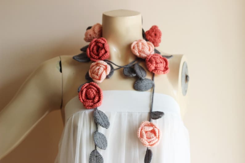 May include: A handmade crochet necklace featuring coral and peach-colored roses with gray leaves. The floral design is draped on a mannequin, showcasing the necklace's length and detail. The necklace is a unique accessory.