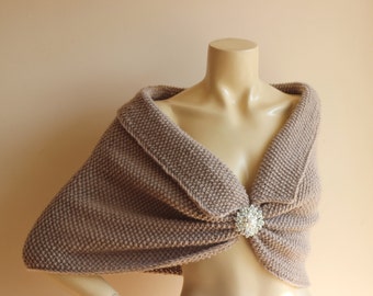 May include: A taupe-colored knitted shawl with a decorative brooch. The shawl is draped over a mannequin, showcasing its collar and draped design. The brooch is a cluster of clear crystals and pearls.